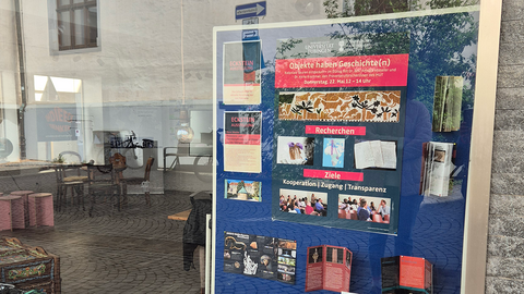 Blick in das Schaufenster des Pop-Up-Stores Eckstein mit einer blauen Stellwand, auf der mittig das Plakat der Veranstaltung "Objekte haben Geschichte(n) – Provenienzforschung am MUT" hängt; links wird im Glas die Straße mit dem Außenbereich eines Cafés reflektiert
