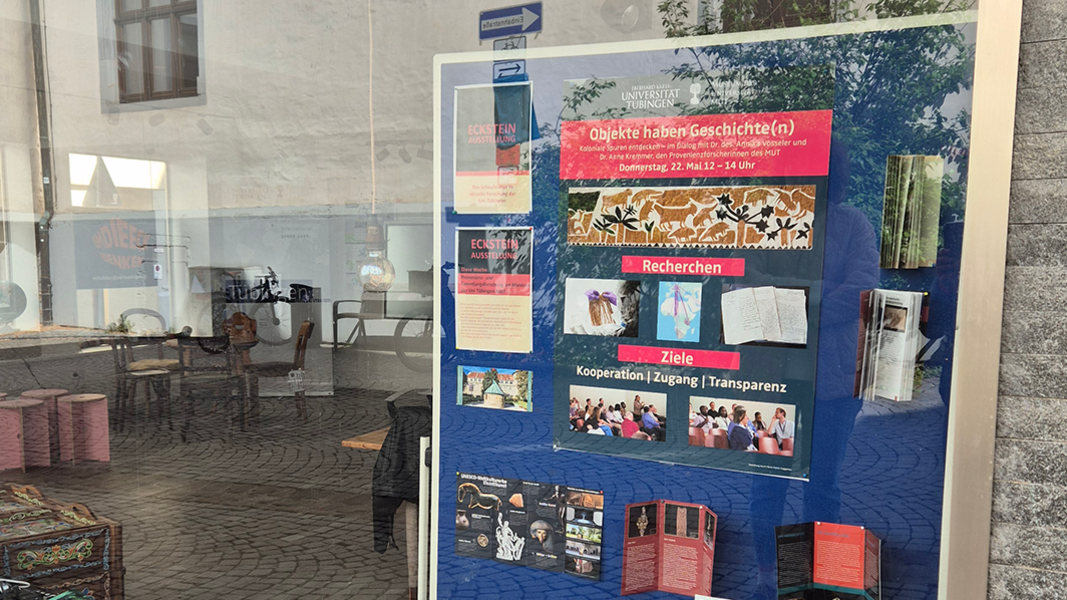 Blick in das Schaufenster des Pop-Up-Stores Eckstein mit einer blauen Stellwand, auf der mittig das Plakat der Veranstaltung "Objekte haben Geschichte(n) – Provenienzforschung am MUT" hängt; links wird im Glas die Straße mit dem Außenbereich eines Cafés reflektiert