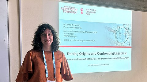Dr. Anne Kremmer steht zur Linken einer Projektionsfläche mit der Titelfolie ihrer Präsentation "Tracing Origins and Confronting Legacies: Provenance Research at the Museum of the University of Tübingen MUT"