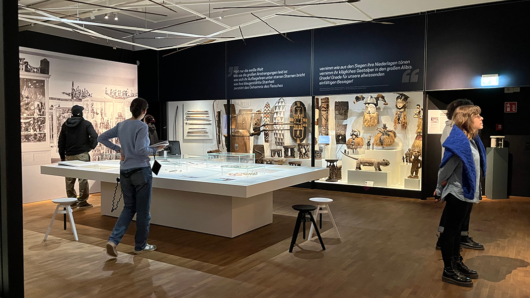 Verschiedene Personen in der Afrika-Abteilung des Linden-Museums mit in die Wand eingelassener Vitrinen mit afrikanischen Waffen und Schilden, in der Mitte ein Tisch mit Informationen und Vitrinenaufbauten