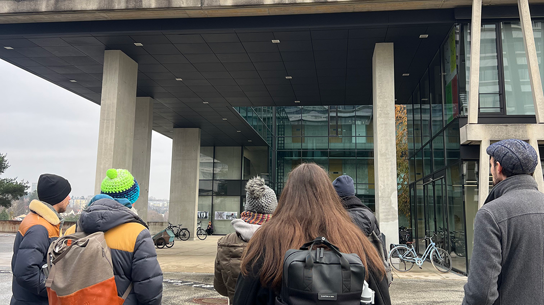 Rückansicht mehrerer Studierender und Dr. Edgar Bierende mit einem Rucksack, die vor einem hohen Vordach mit Betonpfeilern stehen und zur gläsernen Fassade dahinter blicken