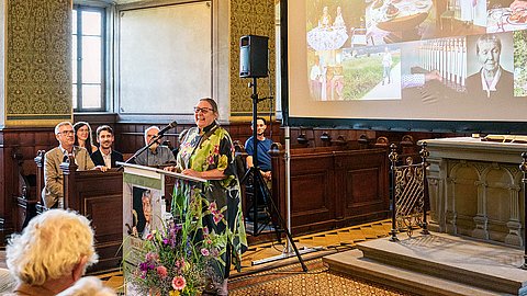 Claudia Schöning-Kalender steht lachend am Redernpult in der Schlosskirche, hinter ihr (rechts) der Altar, vor dem eine Leinwand mit projizierten Bildern aus Evamarie Sanders Leben hängt
