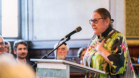 Claudia Schöning-Kalender steht mit ergriffener Geste, die Handfläche auf der Brust, nach links gewandt an einem Rednerpult