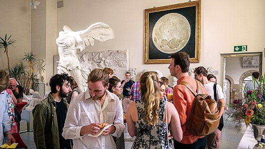 Besuchende der Ausstellungseröffnung stehen in Grüppchen im Rittersaal, links hinter ihnen der Abguss der Nike von Samothrake und mittig das Mondgemälde, im Vordergrund stehen Personen am Buffet