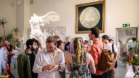 Besuchende der Ausstellungseröffnung stehen in Grüppchen im Rittersaal, links hinter ihnen der Abguss der Nike von Samothrake und mittig das Mondgemälde, im Vordergrund stehen Personen am Buffet