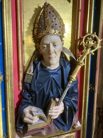 Eine detailreiche Statue eines Abts. Die Figur trägt eine mit Verzierungen versehene Mitra und ein dunkelblaues Gewand. In der rechten Hand hält die Figur ein geöffnetes Buch, in der linken einen verzierten goldenen Bischofsstab. Der Hintergrund ist golden und rot mit blauen Streifen.