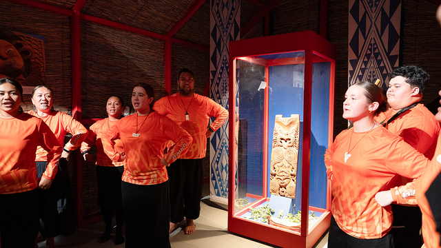 Die Schüler*innen als Vertreter der Te Aitanga-a-Hauiti aus Uawa führen um die beleuchtete Vitrine mit Hinematioros Pou herum einen Tanz auf; alle haben die Hände in die Seiten gestemmt