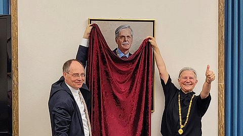 Foto: MUT | Ernst Seidl Zwei Personen halten an je einem Ende ein rotes Samttuch, das die untere Hälfte eines Portraits von Bernd Engler verhüllt