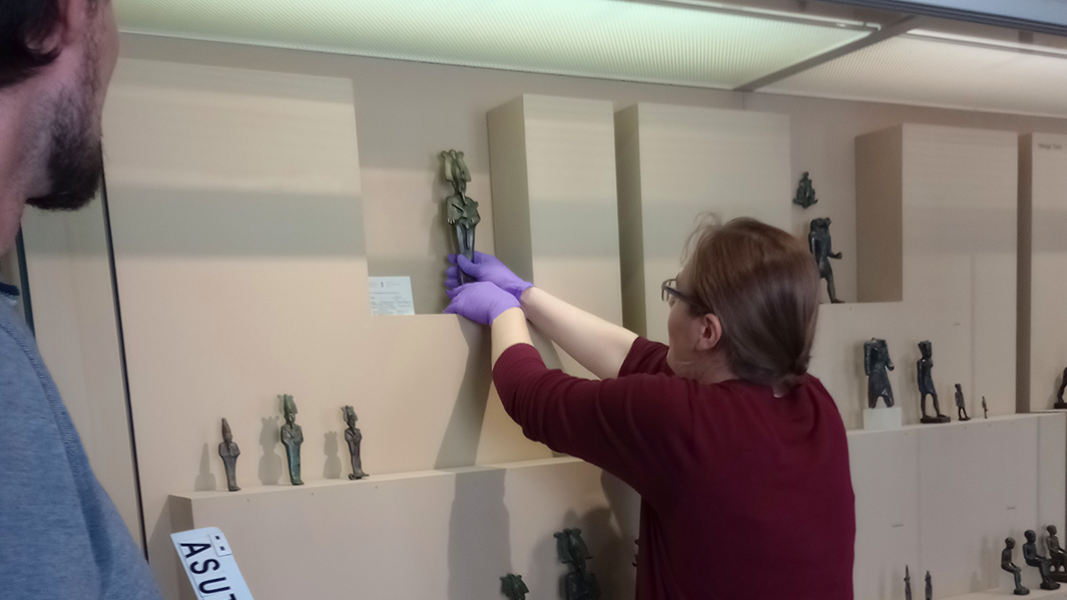 Leah Kupferschmied holt eine Statuette aus der Vitrine mit den Bronzen der Ägyptologischen Sammlung im Museum Alte Kulturen, links im Bild: Fabian Kurze mit der Magnettafel mit den Inventarnummern