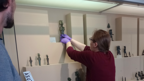 Foto: MUT | Nadja Mozdzen Leah Kupferschmied holt eine Statuette aus der Vitrine mit den Bronzen der Ägyptologischen Sammlung im Museum Alte Kulturen, links im Bild: Fabian Kurze mit der Magnettafel mit den Inventarnummern