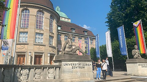 © MUT | Simon Zauner Zu sehen ist der Haupteingang der Universität Zürich mit einigen Flaggen und zwei Mitarbeiterinnen des MUT, Annika Vosseler und Diellëza Hyseni
