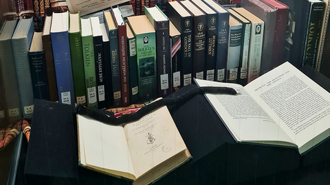 Blick leicht schräg von links in eine hohe Vitrine mit einer Reihe unterschiedlich gestalteter Bücher und zwei aufgeschlagenen Büchern auf Buchstützen davor; darüber hängt mittig ein Schwarz-Weiß-Bild von J. R. R. Tolkien, umgeben von Textausdrucken, die nach oben verbreiternd aufgehangen sind.