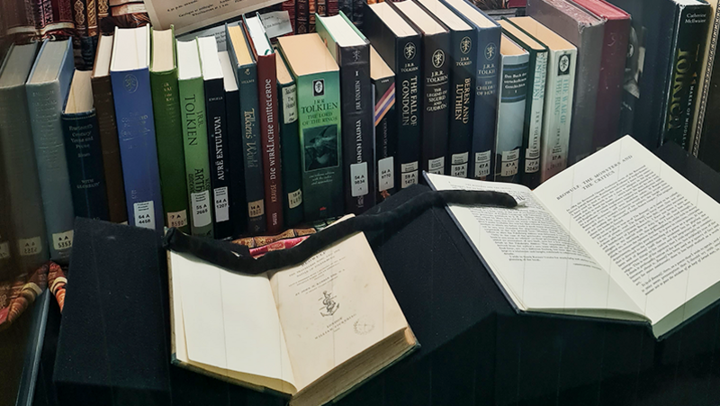 Blick leicht schräg von links in eine hohe Vitrine mit einer Reihe unterschiedlich gestalteter Bücher und zwei aufgeschlagenen Büchern auf Buchstützen davor; darüber hängt mittig ein Schwarz-Weiß-Bild von J. R. R. Tolkien, umgeben von Textausdrucken, die nach oben verbreiternd aufgehangen sind.
