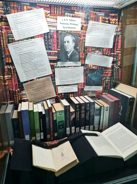 Blick leicht schräg von links in eine hohe Vitrine mit einer Reihe unterschiedlich gestalteter Bücher und zwei aufgeschlagenen Büchern auf Buchstützen davor; darüber hängt mittig ein Schwarz-Weiß-Bild von J. R. R. Tolkien, umgeben von Textausdrucken, die nach oben verbreiternd aufgehangen sind.