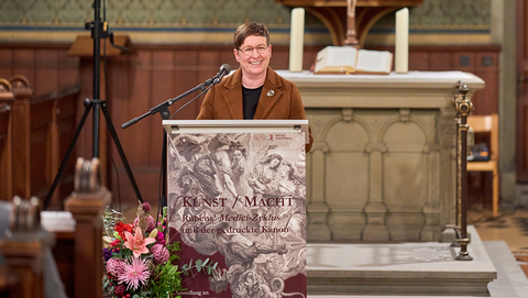 Ariane Koller steht lächelnd hinter dem Rednerpult mit dem Plakat der Ausstellung "Kunst und Macht", im Vordergrund die Ecke der Kirchenbänke der Schlosskirche