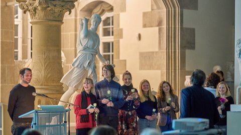 Foto: MUT | Fabian Kurze Am Rednerpult Dr Michael La Corte, daneben (v. l. n. r.) Solveig Schreiber, Timon Butz, Anastasia Rotenberg, Leyla Waberski, Diellëza Hyseni mit jeweils einer weißen Rose in der Hand; ganz rechts im Bild Dr des Annika Vosseler mit Blumenstrauß