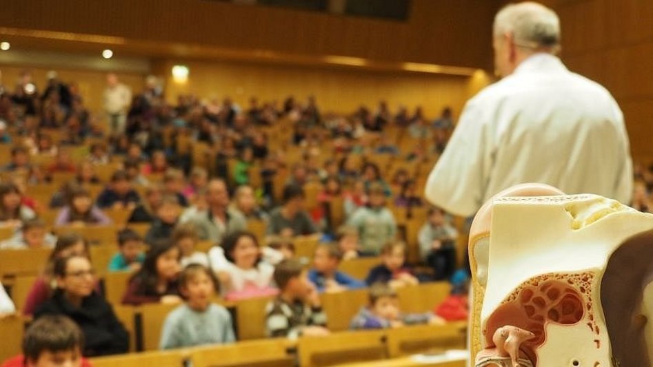 Verschommener Blick auf einen Hörsaal mit Kindern, im Vordergrund scharf ein Dozent im weißen Shirt in der Rückansicht