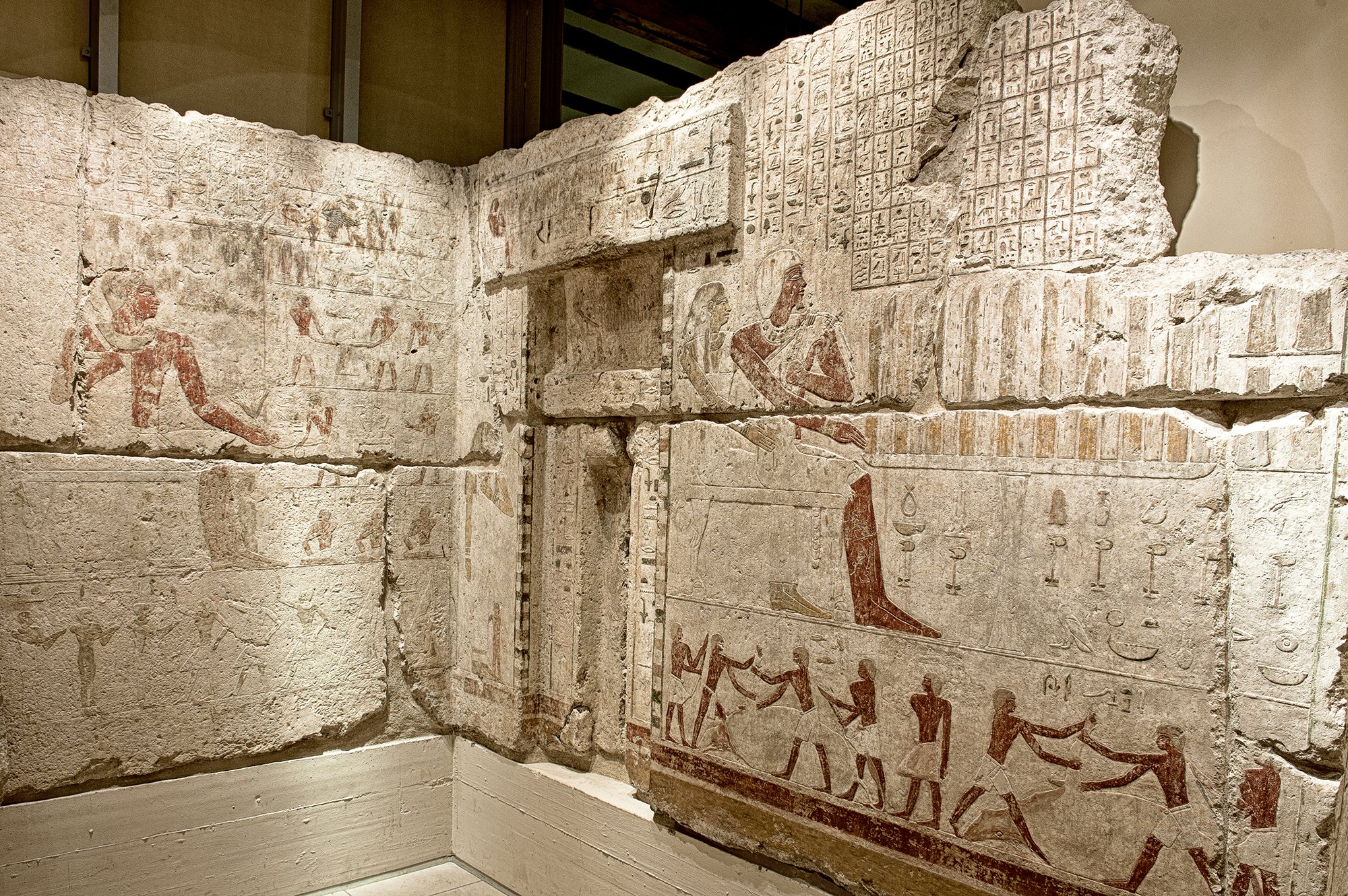 Blick auf zwei im rechten Winkel aufeinandertreffende Wände der ägyptischen Opferkammer mit einer sichtbaren Scheintür; die Wände sind mit hieroglyphischen Inschriften geschmückt, deren Bemalung in Dunkelrot, Beige und Blau sich zum Teil erhalten hat