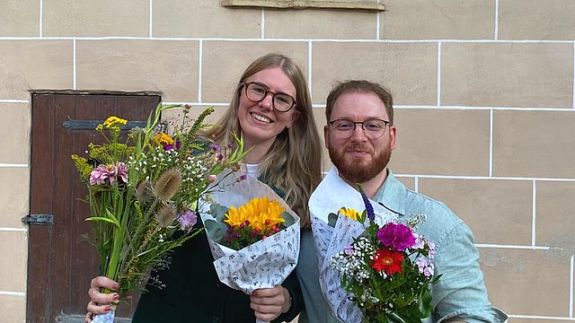 Teresa Pohl (links) und Lucas Rau (rechts) stehen mit bunten Blumensträußen in den Händen vor der gelben Steinmauer des Innenhofs von Schloss Hohentübingen