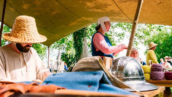Niedriger Blick über einen Tisch mit Textilien, links im Bild ein Mann in weißem Hemd mit ins Gesicht gezogenem Strohhut, rechts und weiter hinten eine Dame mit blauem Kleid und rosa Untergewand mit langem Ärmeln und einer weißen Haube