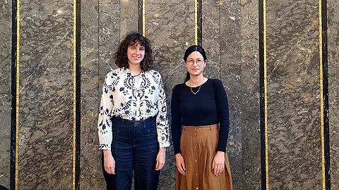 Dr. Anne Kremmer (links) und Jasmin Günther (rechts) vom MARKK stehen vor einer aufrechten Marmorwand mit goldenen und schwarzen Streifen
