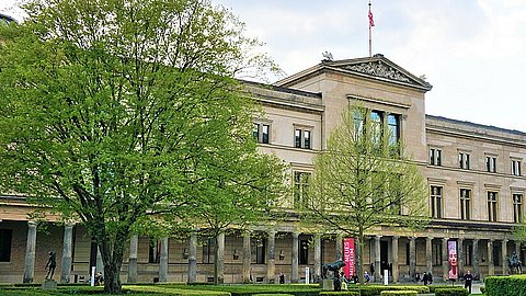 Schräger Blick auf das Neue Museum in Berlin mit palaisartiger Fassade und Arkaden im Erdgeschoss; davor Grasfläche und Begrünung