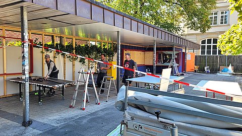 Sommerliche Baustelle des Unipavillons mit Absperrband und einem Ladebett mit dem Schriftzug "Potengowski.com" im Vordergrund, drei Männer stehen an einem Schneidetisch, umgeben von mehreren Leitern