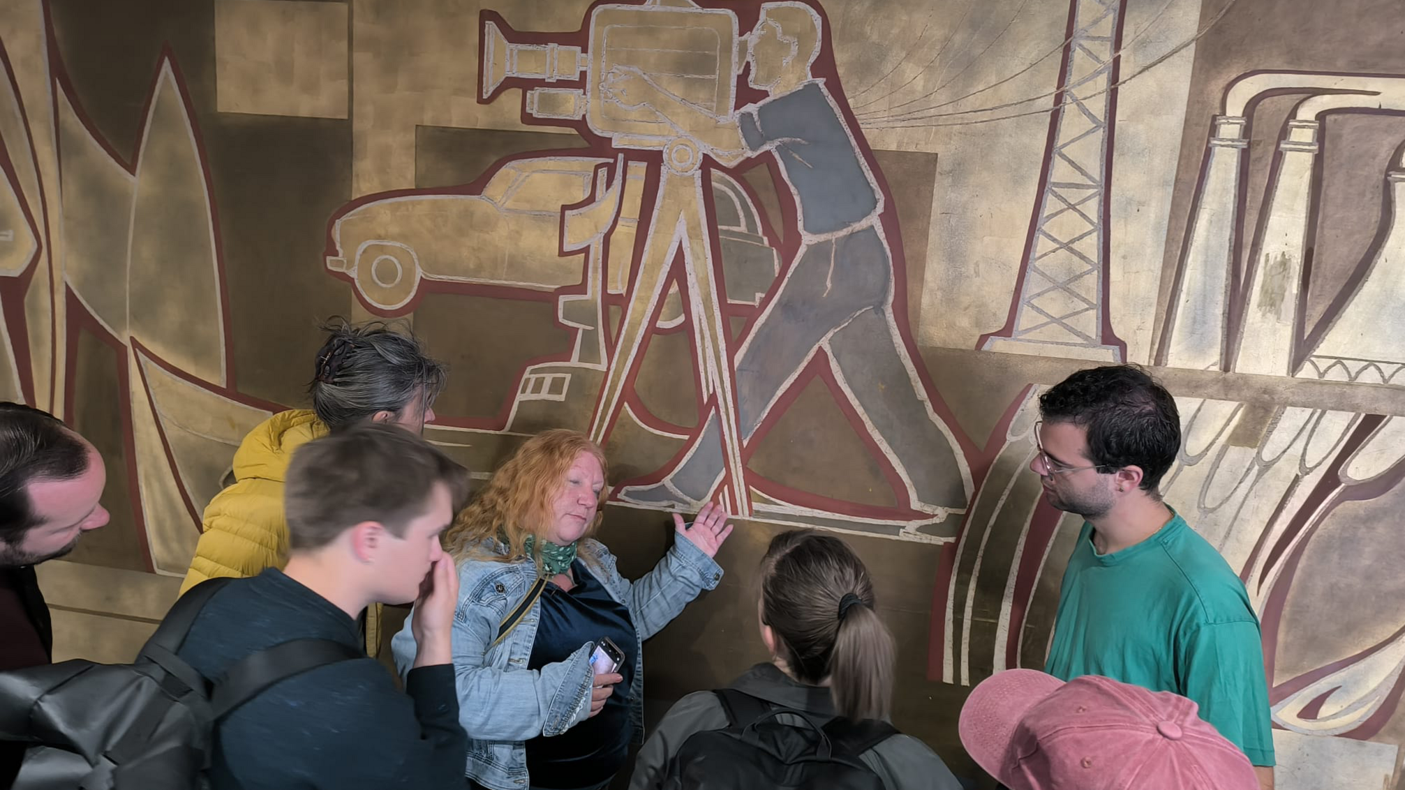 Eine Gruppe schart sich um eine erklärende Person, die auf ein Wandbild mit der Darstellung eines Kameramannes zeigt