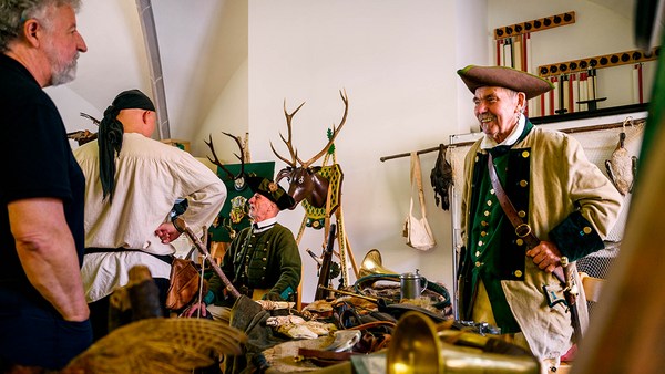 Rechts im Bild ein Wildjäger des 18. Jahrhunderts, umgeben von Jagdinstrumenten und -trophäen, rechts im Bild Besuchende