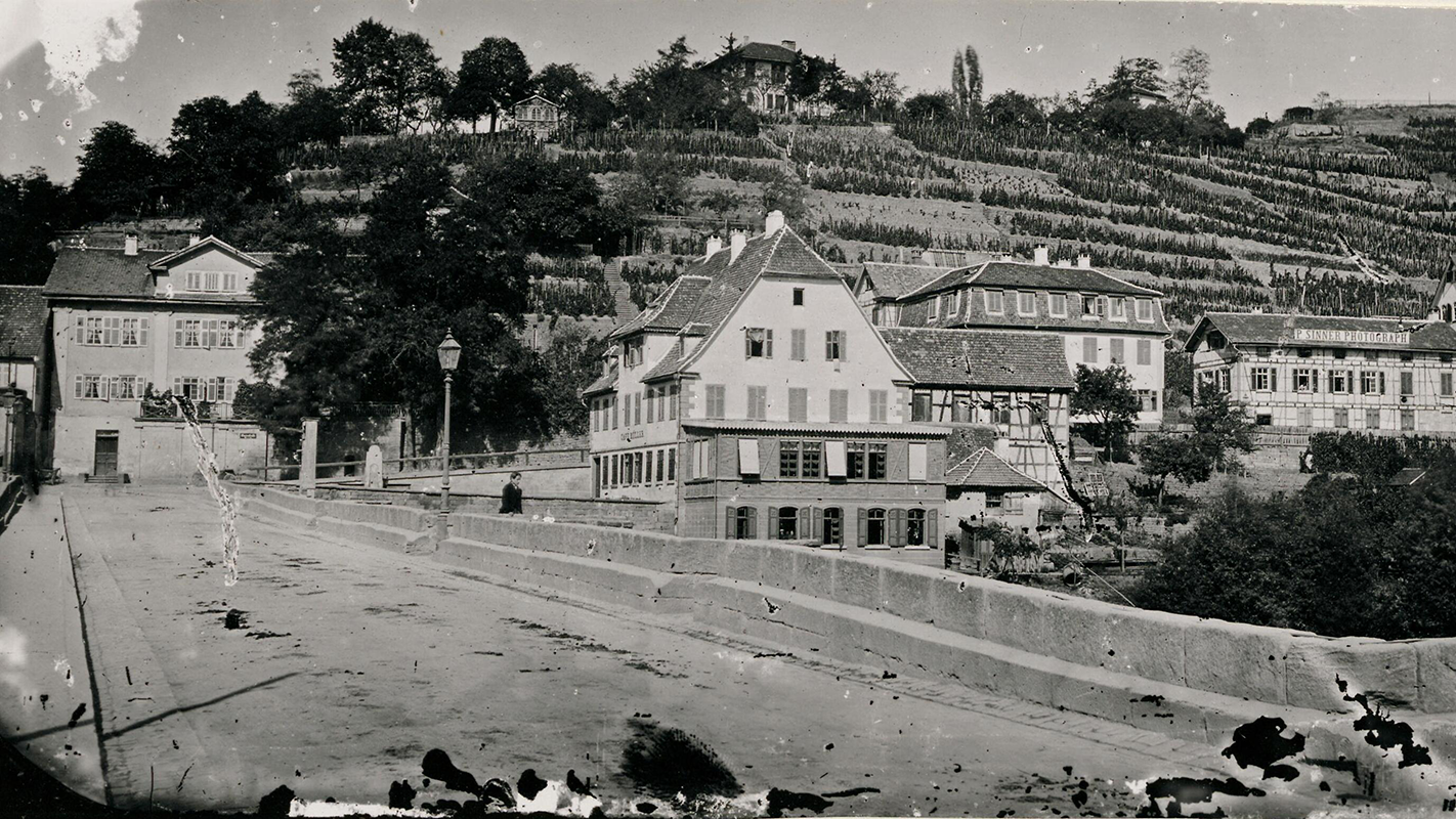 Schwarzweiß-Foto von 1868: Neckarbrücke mit dem 1944 zerstörten Wohnhaus Ludwig Uhlands und  dem Atelier von Paul Sinner in der Gartenstraße 7 mit Werbeschild auf dem Dach.