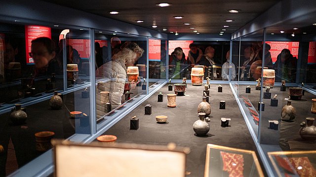 View through the showcase filled with artefacts made from ceramic, jade and other stones; the faces of the surrounding visitors are anonymized through the reflective glass surfaces