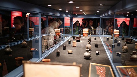 © MUT | Valentin Marquardt Blick längst durch eine lange, beleuchtete Vitrine, die mit Exponaten aus Keramik, Jade und anderem Stein bestückt ist; durch die spiegelnden Glasflächen sind die umstehenden Menschen zu sehen, deren Gesichter verschwommen oder verpixelt sind
