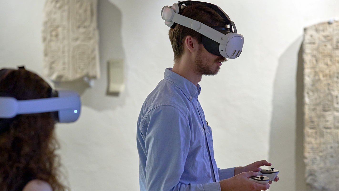 Zu sehen ist der Oberkörper eines nach rechts gewandten Besuchers im blauen Hemd mit weißer VR-Brille, der den Controller der VR-Station hält; im Vordergrund ist unten links unscharf der Kopf einer weiteren Besucherin mit VR-Brille von hinten zu erkennen, im Hintergrund die an der Wand aufgehängten Inschriften in Hieroglyphen-Schrift der Ägyptologie.