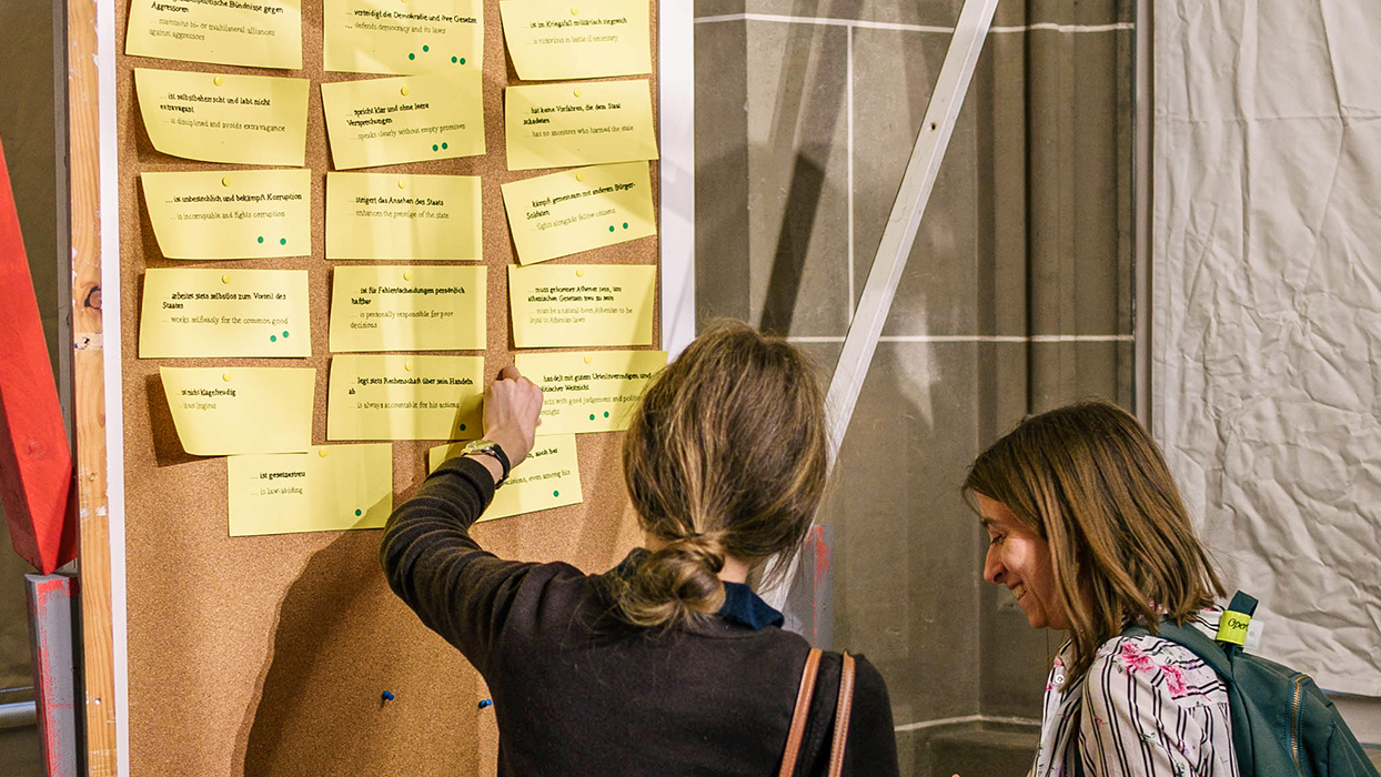 Zwei Besucherinnen stehen vor einer Pinnwand links im Bild, eine zeigt auf einen der gelben Moderationszettel, auf denen Charaktereigenschaften stehen, die einem guten Redner in der Antike zugeschrieben wurden; grüne Punkte markieren vereinzelte Zettel