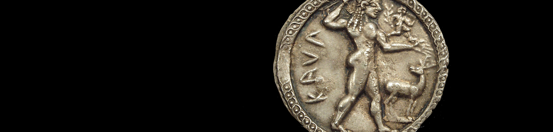 Foto: MUT Bildseite (Avers) einer Silbermünze mit einem jungen, nach rechts schreitenden Apollon, auf dessen ausgestreckten Arm eine kleine, männliche Figur nach rechts rennt; zur Rechten des Gottes wendet sich ein kleiner Hirsch nach ihm um; links des Gottes steht die griechische Inschrift "KAUL" (für Kaulonia)