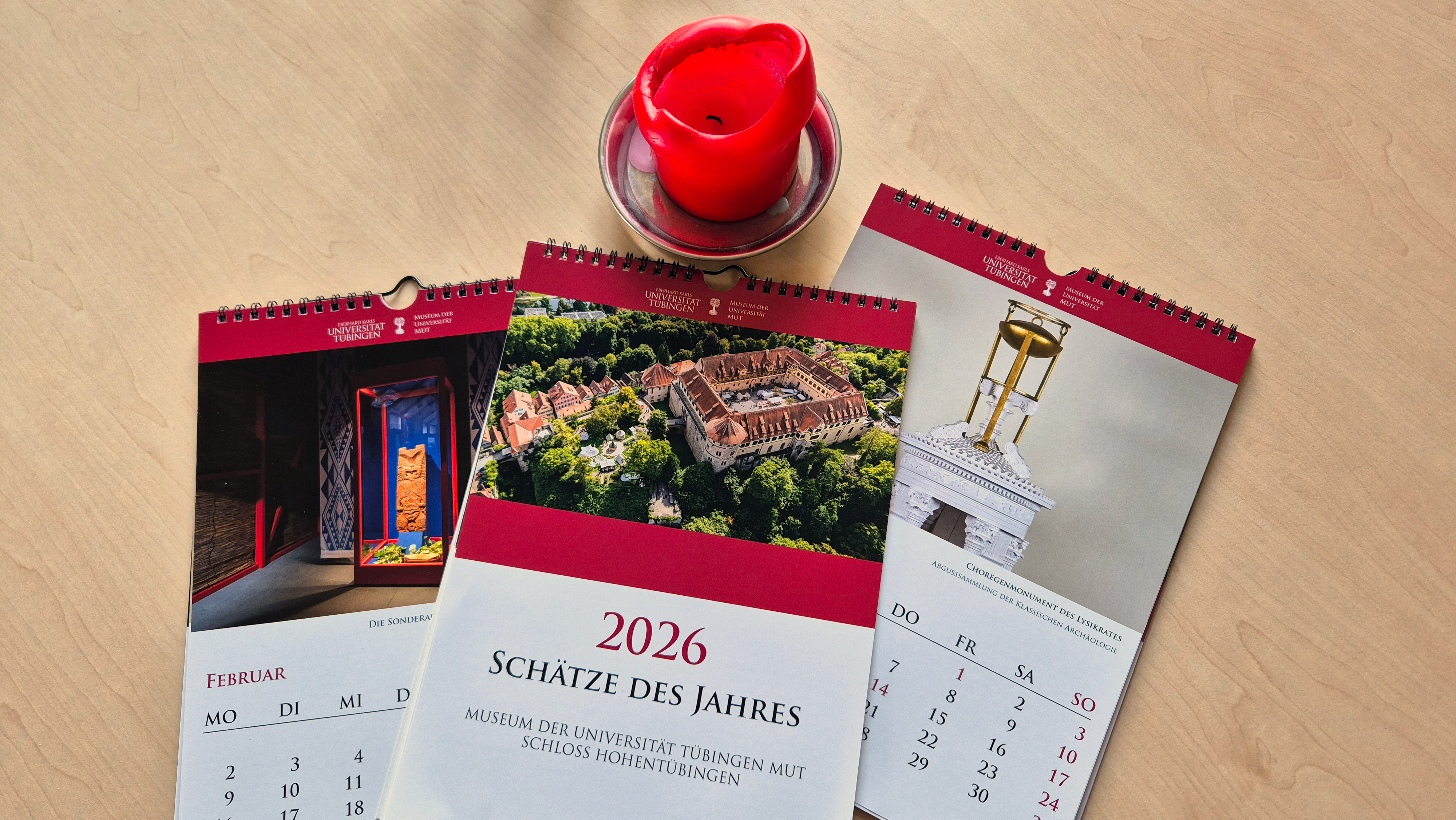 Auf einer Holzplatte liegen drei Jahreskalender neben einer roten Kerze, der mittlere zeigt das Titelblatt "Schätze des Jahres 2026", darunter liegen links und rechts aufgeschlagene Exemplare