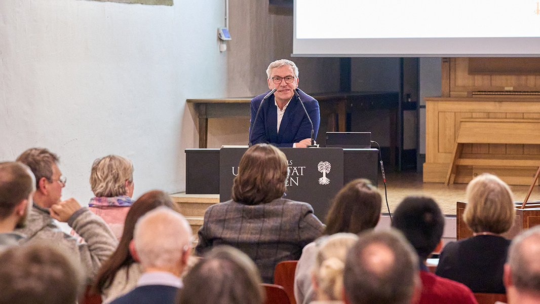 Museumsdirektor Prof. Dr. Ernst Seidl lehnt sich auf das Rednerpult, im Vordergrund die Rückansicht der Köpfe des Publikums