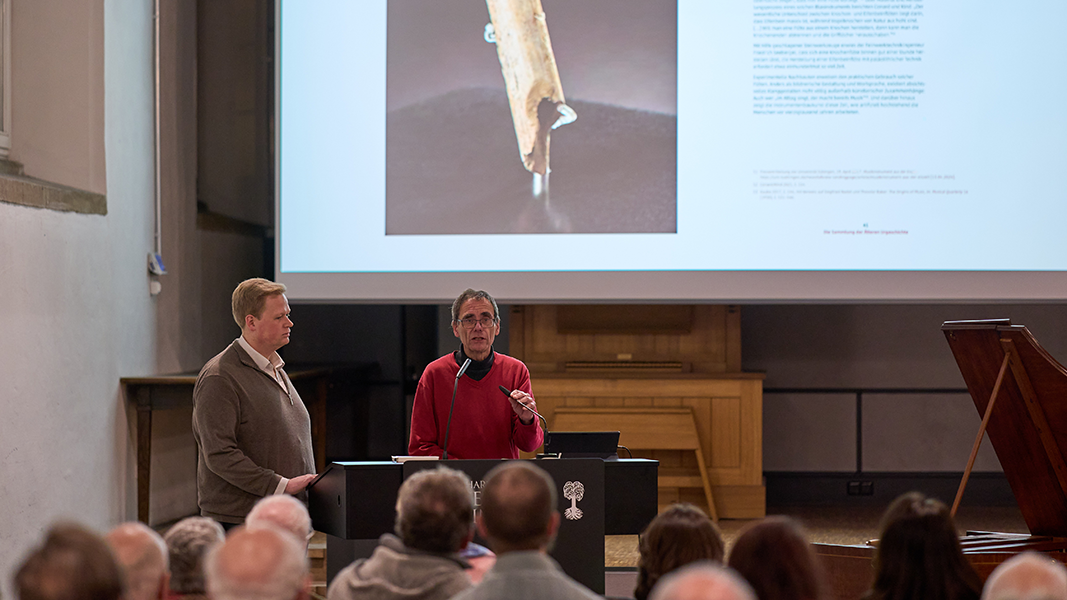 Prof. Dr. Thomas Schipperges und Claudius Hille am Rednerpult, im Hintergrund ist auf der Leinwand die untere Hälfte einer Doppelseite mit dem Bruchstück einer prähistorischen Vogelknochenflöte zu sehen