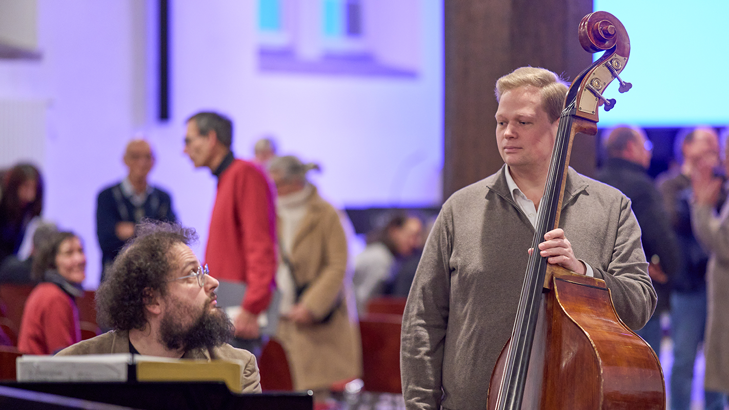Dr. Jörg Büchler am Klavier und Claudius Hille am Cello sehen einander an, im Hintergrund in bläulichem Licht das sich unterhaltende Publikum