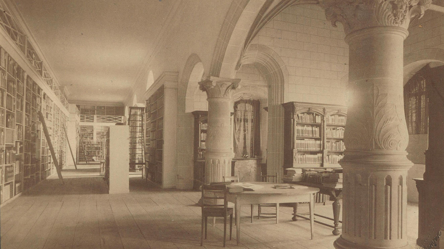 Foto: Universitätsbibliothek Tübingen Schwarzweiß-Foto der Universitätsbibliothek im Rittersaal auf Schloss Hohentübingen mit vollen Bücherregalen an den Wänden und einem Arbeitstisch mit Stühlen zwischen den Kolonnaden